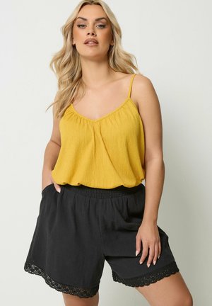 Kvinde med langt blond hår iført en gul ærmeløs top og løse sorte shorts med blonderkant, stående med den ene hånd i lommen.