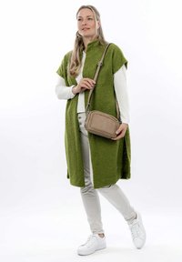 Donna che indossa un lungo gilet verde textured sopra una camicia bianca e pantaloni beige, che tiene una borsa a tracolla beige, in piedi con le gambe incrociate.