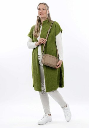 Donna che indossa un lungo gilet verde textured sopra una camicia bianca e pantaloni beige, che tiene una borsa a tracolla beige, in piedi con le gambe incrociate.