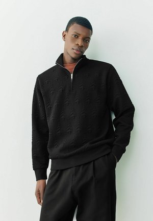 Schwarzer Quarter-Zip-Sweatshirt mit strukturiertem Baum-Muster, gerippten Bündchen und Saum, kombiniert mit einem rostfarbenen Shirt und dunklen Hosen.