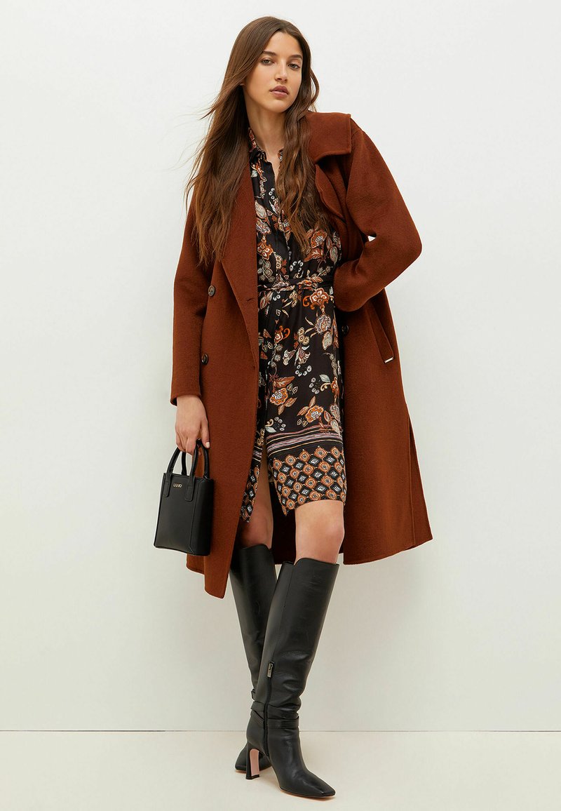 Cappotto di lana marrone sopra un vestito floreale nero con accenti arancioni, abbinato a stivali di pelle nera al ginocchio e una piccola borsa nera.