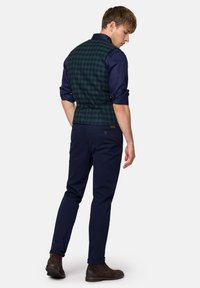 Zielono-granatowa kamizelka w kratę noszona na ciemnoniebieskiej koszuli, zestawiona z granatowymi spodniami i ciemnymi butami, prezentująca dopasowany krój i gładką teksturę.