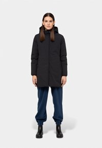 Piumino nero imbottito con cappuccio, dotato di cerniera a tutta lunghezza e tasche laterali, indossato sopra pantaloni da jogging blu scuro e stivaletti neri alla caviglia.