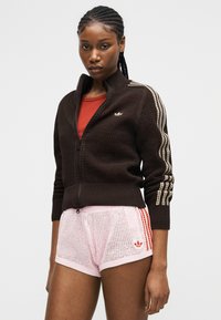 Frau trägt schwarzen Reißverschluss-Pullover mit beigen Streifen an den Ärmeln und hellrosa Mesh-Shorts mit roten Streifen und Logo, steht vor weißem Hintergrund.