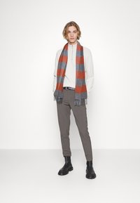 Grå og oransje rutete skjerf, blanding av ull, med franske kanter. Modellen har på seg en lys genser, mørke bukser og svarte støvler. Enkelt, moderne design.