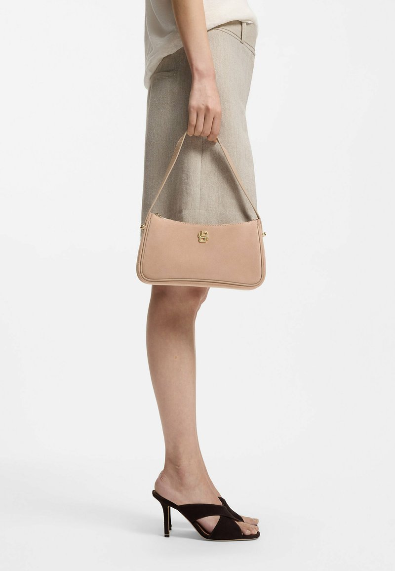 Femme tenant un sac à bandoulière en cuir nude, portant une jupe beige et des sandales à talons noires ouvertes, se tenant devant un fond blanc uni.