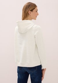 Donna di profilo che indossa una felpa bianca con cappuccio e jeans blu su uno sfondo rosa chiaro.