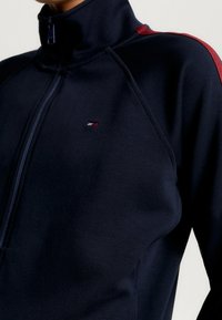 Giacca zip-up navy con colletto alto, realizzata in tessuto liscio. Presenta dettagli rossi sulle spalle e un piccolo logo sul petto.
