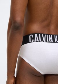 Λευκά βαμβακερά σλιπάκια με μαύρη ελαστική μέση που φέ�ρει την επιγραφή "CALVIN KLEIN" σε λευκά γράμματα, παρουσιάζοντας λεία υφή και εφαρμοστό σχέδιο.