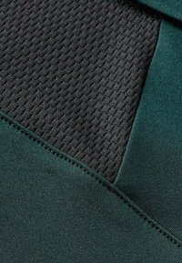 Tejido deportivo de color verde oscuro con un inserto de malla negra texturizada. Textura suave con materiales contrastantes y finos detalles de costura.