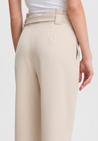 Pantaloni beige a vita alta con taglio sartoriale, dotati di tasche laterali e una texture liscia. Passanti per la cintura visibili in vita.