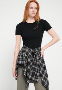 Black fitted t-shirt paired with a black and grey plaid shirt tied around the waist. Model wears olive-green fitted pants.