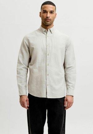 Homme portant une chemise beige clair à manches longues boutonnée et un pantalon noir, debout devant un fond blanc uni.