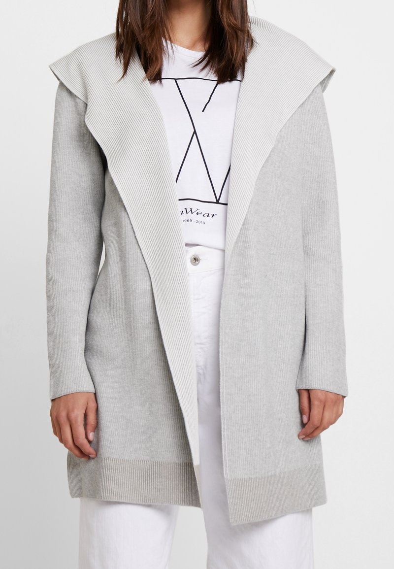 Femme portant un long cardigan en maille gris clair à larges revers, par-dessus un t-shirt blanc à motif et un pantalon blanc, debout devant un fond uni.