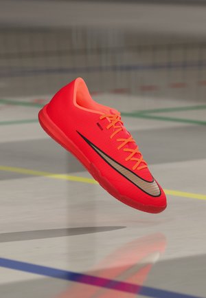 Jasnoczerwony halowy but do piłki nożnej z czarno-złotym logo Nike, pomarańczowe sznurowadła, unoszący się nad podłogą sali gimnastycznej z liniami boiska.