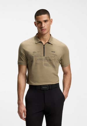 Uomo che indossa una polo beige a maniche corte con una zip nera e pantaloni neri, con le mani nelle tasche, su sfondo bianco neutro.