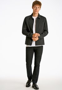 Mörkgrå knäppt jacka med krage och bröstficka, över en vit t-shirt, i kombination med svarta slim fit byxor och svarta skor.