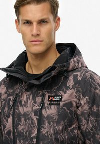 Man in een zwart en bruin gemêleerd Superdry Mountain hooded jack met ritszakken, recht naar voren kijkend tegen een effen achtergrond.