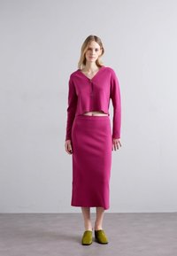 Fuchsia cropped cardigan over a matching fitted pencil skirt. The outfit has a smooth texture and is completed with green heeled shoes.