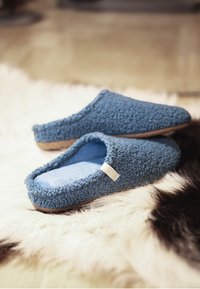 Pair of blue textured slip-on slippers resting on a white and dark brown fuzzy rug on a hardwood floor.