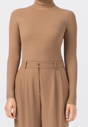 Kvinde iført en tætsiddende ribbet kamelrullekravesweater, stukket ned i højtaljede, vidde kamelbukser med bæltestropper.