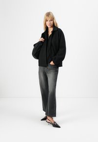 Kvinne med svart jakke, svart topp, grå vide benjeans, svarte slingback-hels, holder svart veske over skulderen mot hvit bakgrunn.