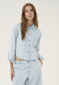 Light blue denim cropped jacket with a button-up front, chest pocket, and slightly rolled sleeves. Soft fabric with a smooth texture.