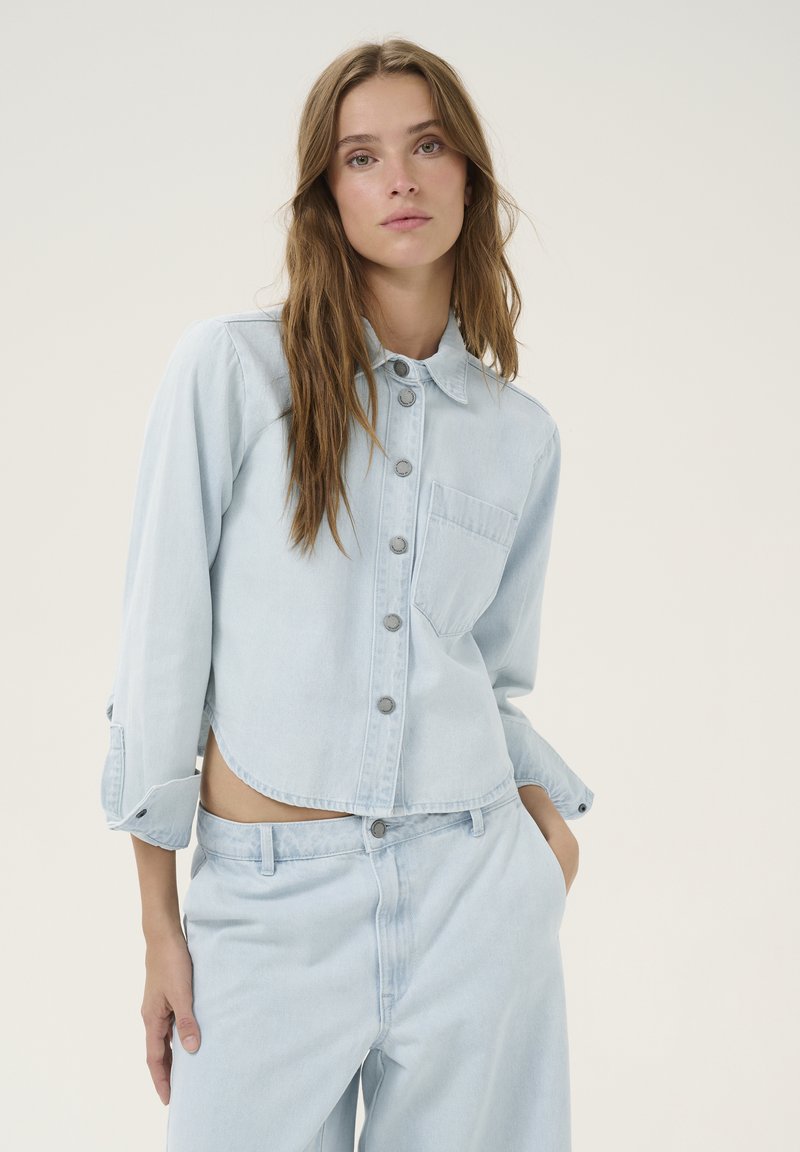 Light blue denim cropped jacket with a button-up front, chest pocket, and slightly rolled sleeves. Soft fabric with a smooth texture.