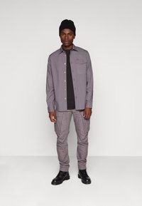Camisa de botones gris con un bolsillo en el pecho, usada sobre una camiseta negra, combinada con pantalones cargo gris claro y botas negras con cordones. Gorro negro.