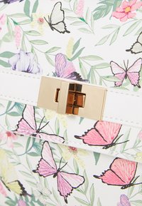 Sac à main à motif floral sur un fond blanc, décoré de papillons colorés et équipé d'un fermoir en métal doré sur une surface texturée.