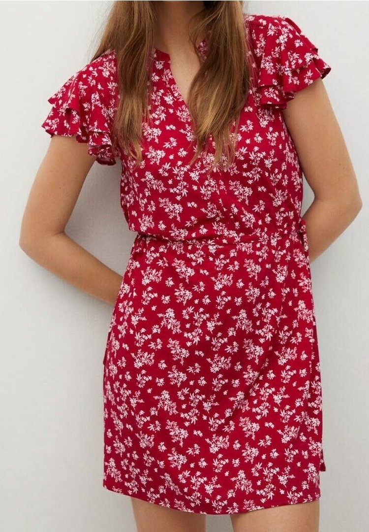 Femme portant une robe courte rouge avec un motif floral blanc et des manches courtes à volants, debout les mains dans le dos devant un fond uni.