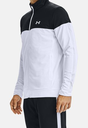 Man draagt een zwart-wit sportjack met rits en het Under Armour-logo op de borst, gecombineerd met een zwarte broek met witte strepen.