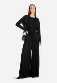 Schwarze Strickjacke mit Knopfleiste, kombiniert mit hochgeschnittenen, schwarz-grün gestreiften weiten Hosen. Akzentuiert mit spitzen, genagelten Schuhen.