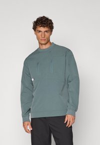 Genser i dempet teal, med høy krage, to brystlommer med glidelås og ribbestrikkede mansjetter. Stoffet har en glatt, matt tekstur.