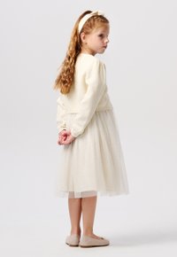 Pull en tricot blanc et robe en tulle, tous deux avec une texture douce. La robe présente un voile transparent avec des paillettes. Ballerines de couleur claire.