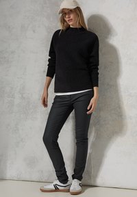 Jersey negro de punto con cuello alto, combinado con pantalones oscuros ajustados y zapatillas blancas con rayas negras. La gorra completa el look.