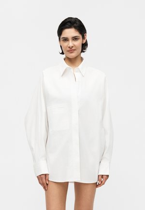 Femeie cu păr scurt și închis la culoare, purtând o cămașă albă supradimensionată cu nasturi și mâneci lungi, stând în fața unui fundal simplu, deschis.