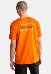 Hombre con cabeza rapada que lleva una camiseta naranja de Timberland con el texto "Año del Tigre" y "La naturaleza necesita héroes" en la espalda.