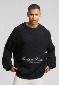 Maglione nero lavorato a maglia con scollo rotondo, vestibilità ampia e polsini a costine. Presenta la scritta bianca "Another Cotton Established 2023" sul davanti.
