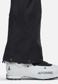 Zwarte, waterdichte skibroek met drukknoopsluitingen aan de onderkant, gecombineerd met een witte skischoen met een grijs "ATOMIC" logo en blauwe accenten.