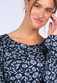 Junge Frau mit hellbraunem Haar, die ein marineblaues, blumiges Langarmshirt trägt und sanft lächelt, während eine Hand nah an ihrem Gesicht ist.