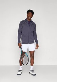 Mannen atletische outfit met een marineblauwe ritsvest, witte korte broek en tennisschoenen. Het vest heeft een gladde textuur en een getailleerd ontwerp.