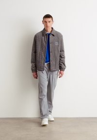 Chaqueta de pana gris con botones a presión, con un logo bordado. Estilo sobre un sudadera azul y pantalones grises, combinada con zapatillas blancas.