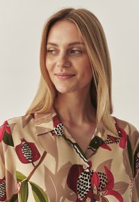 Jonge vrouw met lang blond haar, gekleed in een beige shirt met rode, groene en zwarte bloemenpatronen, die iets opzij kijkt en glimlacht.