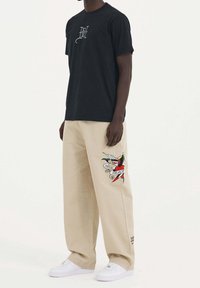 Homme portant une chemise noire à manches courtes et un pantalon beige à jambes larges avec un graphisme coloré style tatouage sur la cuisse droite, et des baskets blanches.