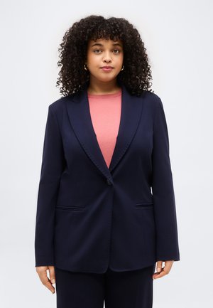 Kobieta o kręconych włosach, nosząca złote kolczyki koła, granatową marynarkę zapinaną na guziki na różowy top, stojąca na jednolitym jasnym tle.