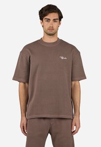 Brun ribbet kortærmet t-shirt med rund hals, der har et hvidt logo på venstre side. Materialet ser ud til at være struktureret og blødt.