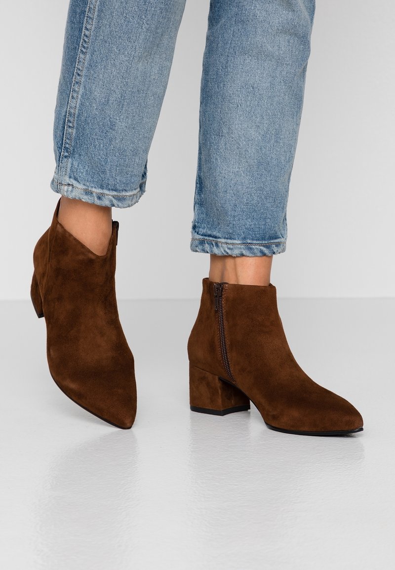 Bottes chevilles en daim marron avec talons carrés portées avec un jean bleu clair retroussé sur un fond clair uni.