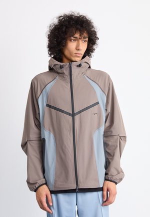 Lichtgewicht bruin jack met blauwe en grijze accenten, capuchon en zwarte rits. Beschikt over elastische manchetten en zijzakken met een slank design.