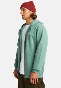 Felpa verde menta con zip e due tasche frontali, cappuccio regolabile con lacci e polsini a coste. Abbinata a una maglietta bianca e pantaloni marroni.